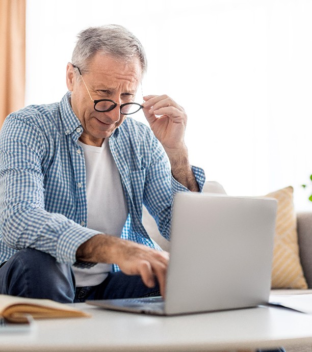 Man on couch lowering glasses and squinting at laptop